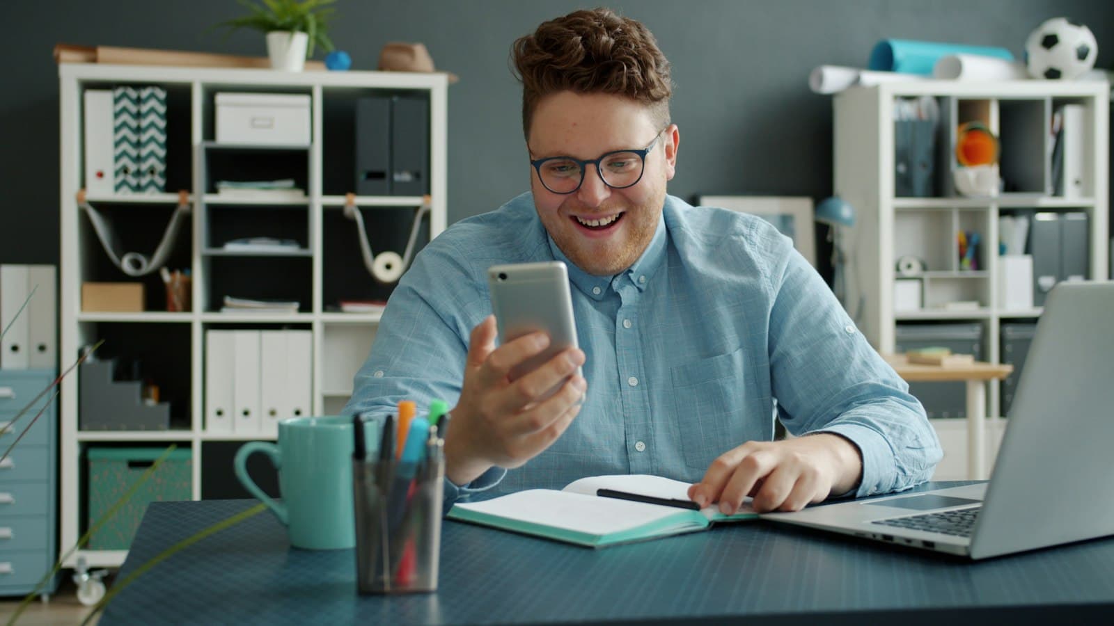 A smiling professional at a desk checking a message on his phone with a notebook and laptop open.